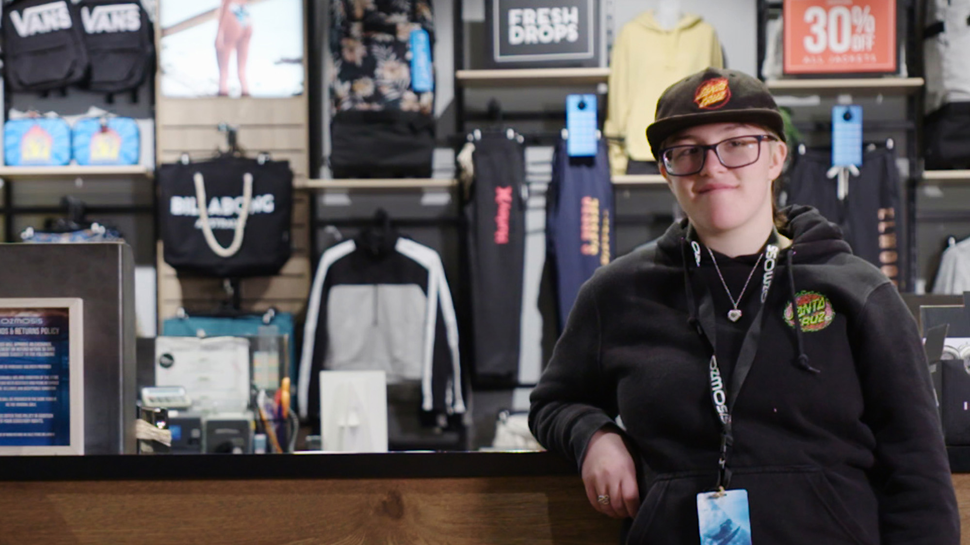 Danielle leaning on Ozmosis counter, wearing ozmosis clothes and has a lanyard around neck